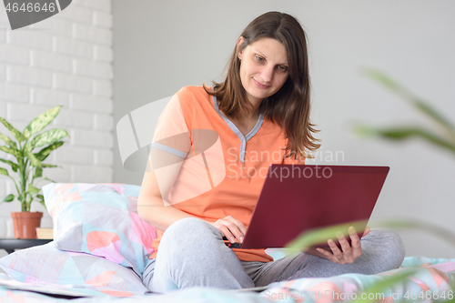 Image of The girl in pajamas works in the computer sitting at home on the bed