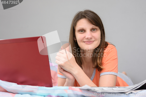Image of The girl lies on the bed with a laptop and a newspaper and looks joyfully into the frame