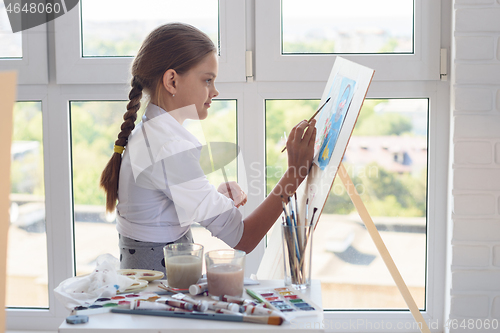 Image of Young artist paints watercolor paints on an easel against the background of a large window