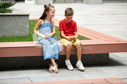 Image of Two smiling kids, boy and girl playing smartphone together in town, city in summer day