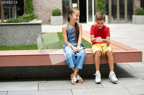Image of Two smiling kids, boy and girl playing smartphone together in town, city in summer day