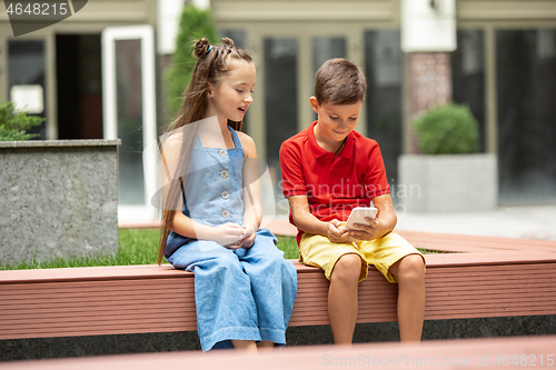 Image of Two smiling kids, boy and girl playing smartphone together in town, city in summer day