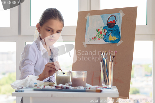 Image of girl sits behind an easel and washes her brush in a glass of water