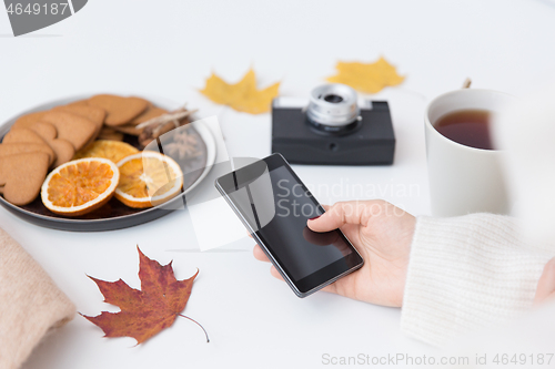 Image of hand with smartphone, tea and autumn leaves