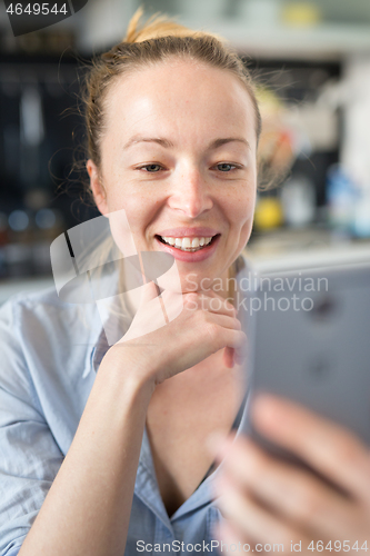 Image of Young smiling cheerful pleased woman indoors at home kitchen using social media apps on mobile phone for chatting and stying connected with her loved ones. Stay at home, social distancing lifestyle.