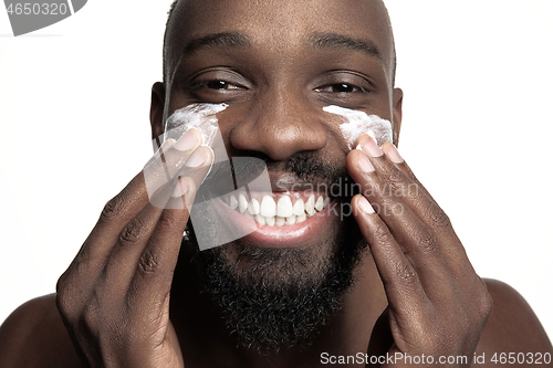 Image of Young african-american guy applying face cream under his eyes on white background