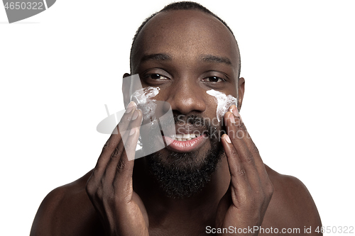 Image of Young african-american guy applying face cream under his eyes on white background