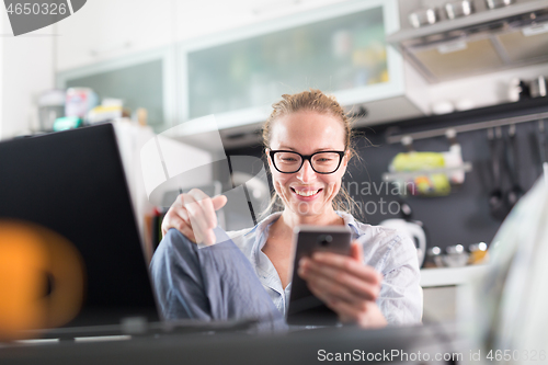 Image of Stay at home and social distancing. Woman in her casual home clothing working remotly from kitchen dining table. Video chatting using social media with friend, family, business clients or partners