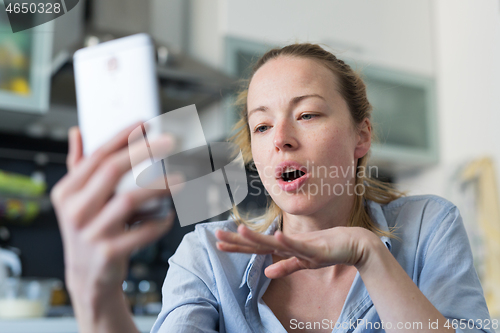 Image of Young pleased woman indoors at home kitchen using social media apps on mobile phone for chatting and stying connected with her loved ones. Stay at home, social distancing lifestyle.