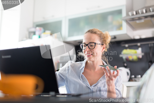 Image of Stay at home and social distancing. Woman in her casual home clothing working remotly from kitchen dining table. Video chatting using social media with friend, family, business clients or partners