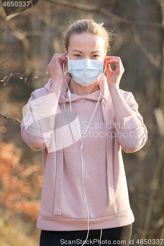 Image of Portrait of caucasian sporty woman wearing medical protection face mask while walking in park, relaxing and listening to music. Corona virus, or Covid-19, is spreading all over the world