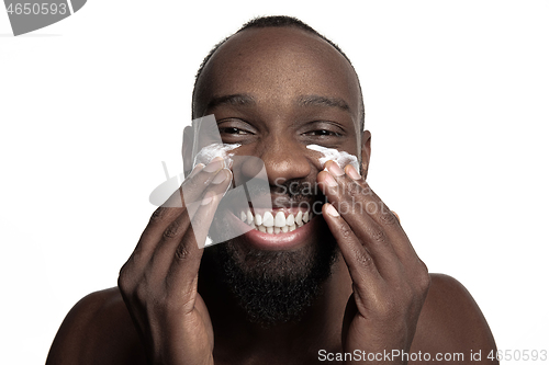 Image of Young african-american guy applying face cream under his eyes on white background