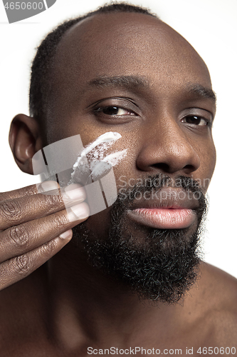 Image of Young african-american guy applying face cream under his eyes on white background