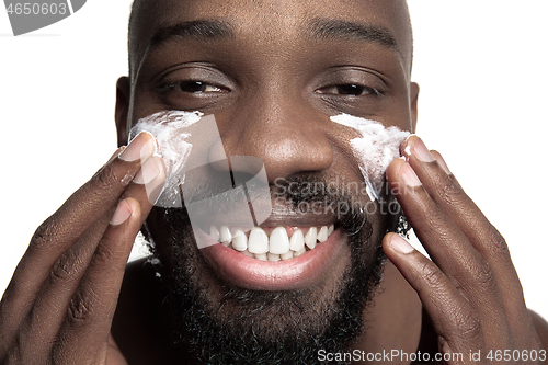 Image of Young african-american guy applying face cream under his eyes on white background