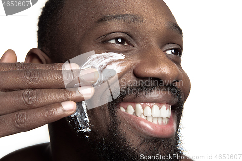 Image of Young african-american guy applying face cream under his eyes on white background