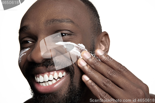 Image of Young african-american guy applying face cream under his eyes on white background