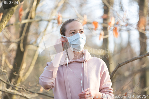 Image of Portrait of caucasian sporty woman wearing medical protection face mask while walking in park, relaxing and listening to music. Corona virus, or Covid-19, is spreading all over the world