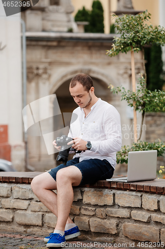 Image of Young photographer with laptopon stone bench andtaking photo on camera