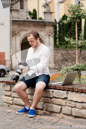 Image of Young photographer with laptopon stone bench andtaking photo on camera