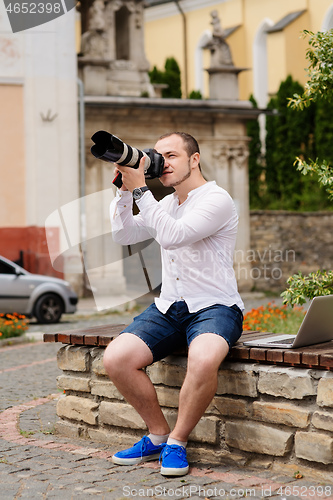 Image of Young photographer with laptopon stone bench andtaking photo on camera