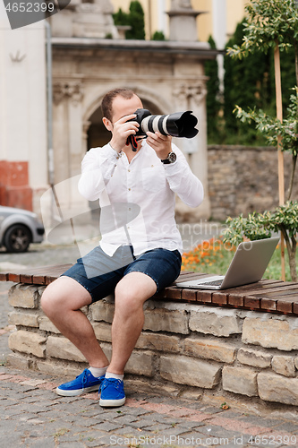 Image of Young photographer with laptopon stone bench andtaking photo on camera