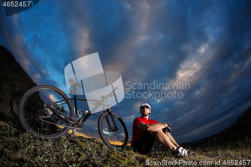 Image of Cyclist resting on grass in mountains. Man is looking aside.