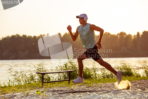 Image of A young athletic man working out listening to the music at the riverside outdoors