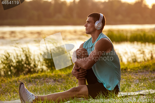 Image of A young athletic man working out listening to the music at the riverside outdoors