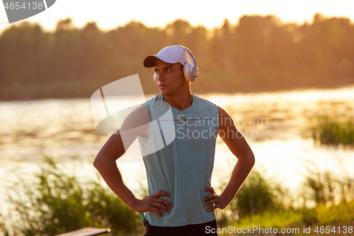 Image of A young athletic man working out listening to the music at the riverside outdoors