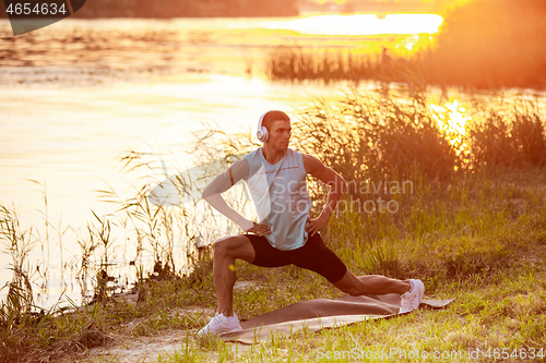 Image of A young athletic man working out listening to the music at the riverside outdoors