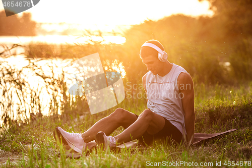 Image of A young athletic man working out listening to the music at the riverside outdoors