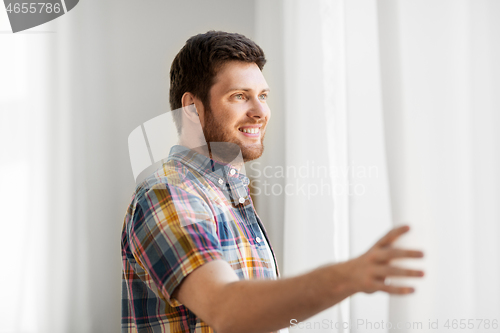 Image of young man opening window curtain at home