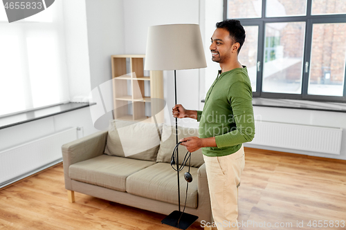 Image of happy indian man holding floor lamp at home