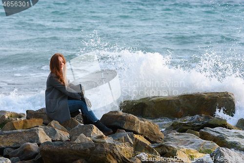 Image of Girl in autumn windy weather sits by the sea on a rocky shore