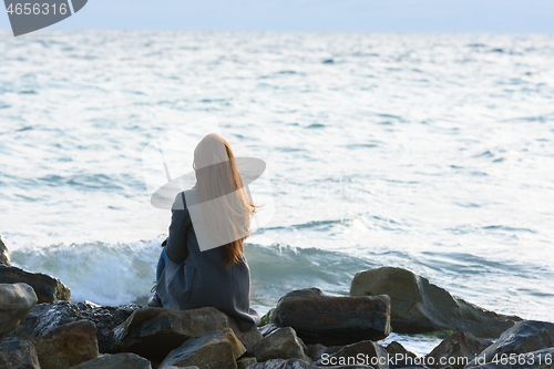 Image of A lonely girl sits on the rocks by the sea and looks into the distance