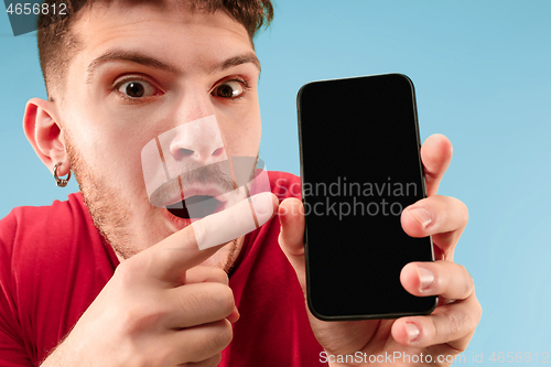 Image of Young handsome man showing smartphone screen isolated on blue background in shock with a surprise face