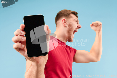 Image of Young handsome man showing smartphone screen isolated on blue background in shock with a surprise face