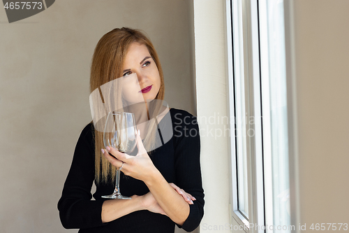 Image of The girl stands thoughtfully at the window with a glass of red wine