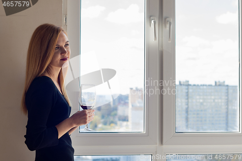 Image of Beautiful brooding girl with a glass of red wine stands near the window