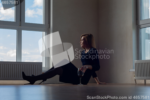 Image of girl in evening dress and shoes sits on the floor near a large window with wine