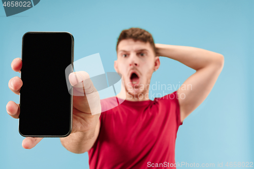 Image of Young handsome man showing smartphone screen isolated on blue background in shock with a surprise face