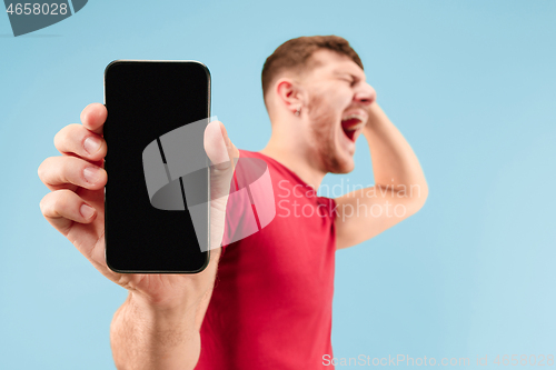 Image of Young handsome man showing smartphone screen isolated on blue background in shock with a surprise face