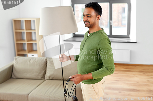 Image of happy indian man holding floor lamp at home