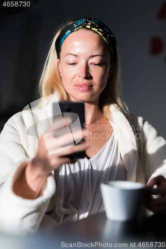 Image of Beautiful caucasian woman at home, feeling comfortable wearing white bathrobe, taking some time to herself, drinking morning coffee and reading news on mobile phone device in the morning