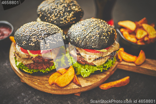 Image of Homemade delicious black burgers served on wooden cutting board with ketchup, potato wedges