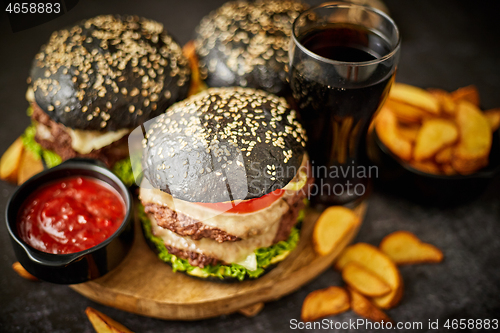 Image of Homemade delicious black burgers served on wooden cutting board with ketchup, potato wedges