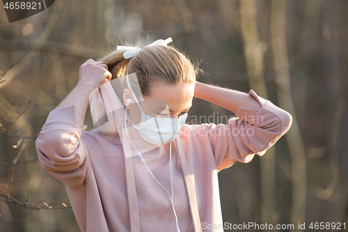 Image of Portrait of caucasian sporty woman wearing medical protection face mask while walking in park, relaxing and listening to music. Corona virus, or Covid-19, is spreading all over the world