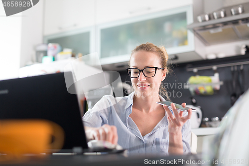 Image of Stay at home and social distancing. Woman in her casual home clothing working remotly from kitchen dining table. Video chatting using social media with friend, family, business clients or partners