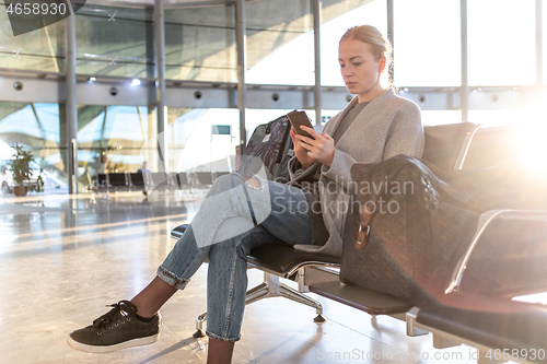 Image of Casual blond young woman using her cell phone while waiting to board a plane at airport departure gates. Empty airport terminal due to corona virus pandemic