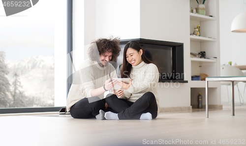 Image of multiethnic couple using tablet computer in front of fireplace